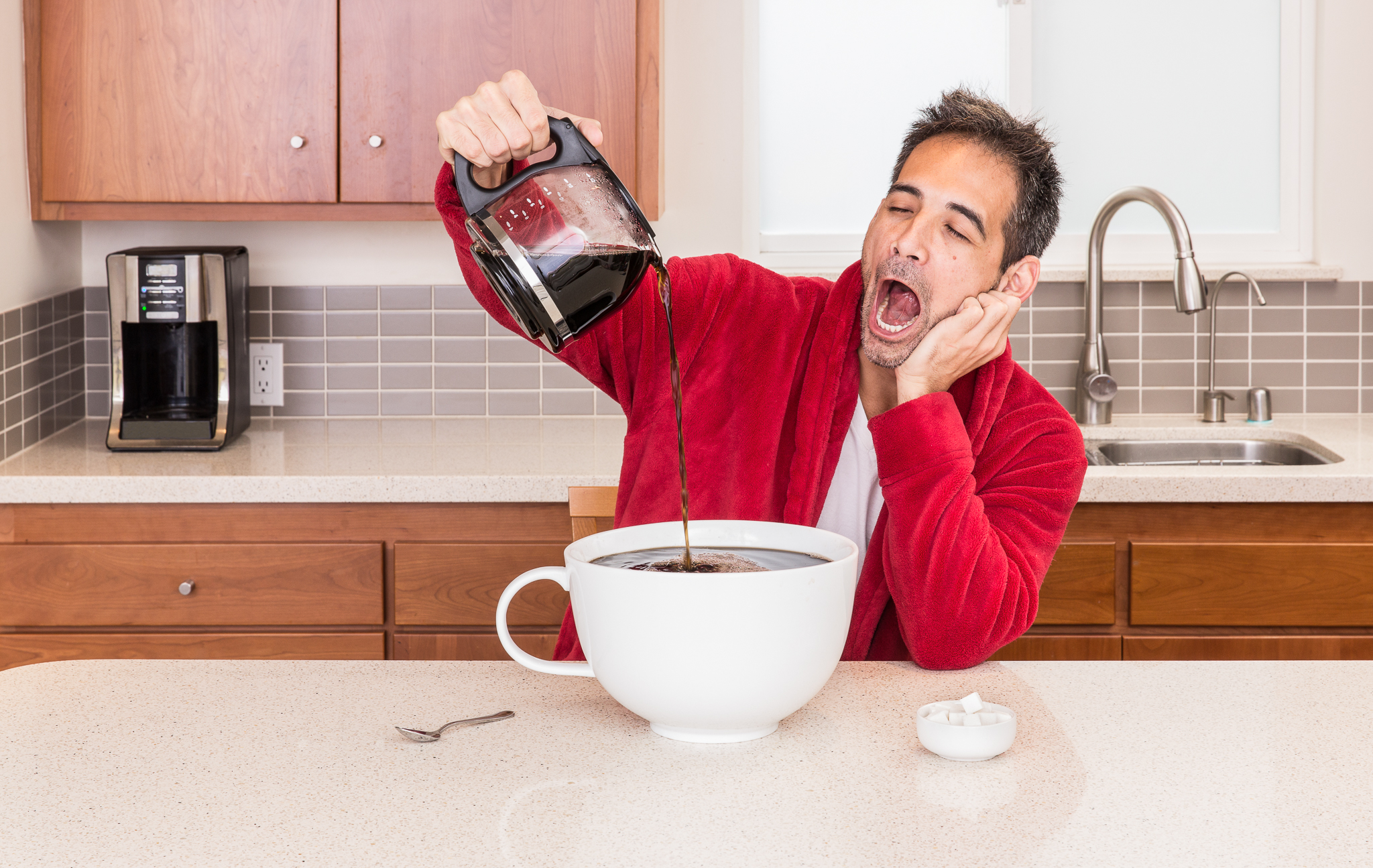 Yawning man pouring coffee into very large coffee cup at kitchen counter.