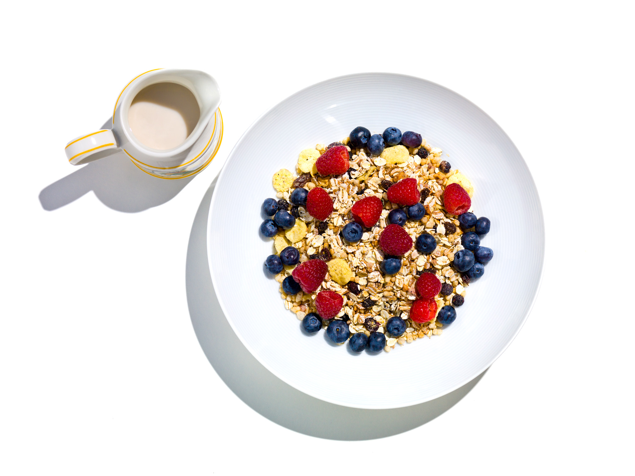 Photograph of a white bowl of granola topped with raspberries and blueberries on a white background with a pitcher of milk next to the bowl. for New York Times - Magazine © 2011 Dana Hursey Photography