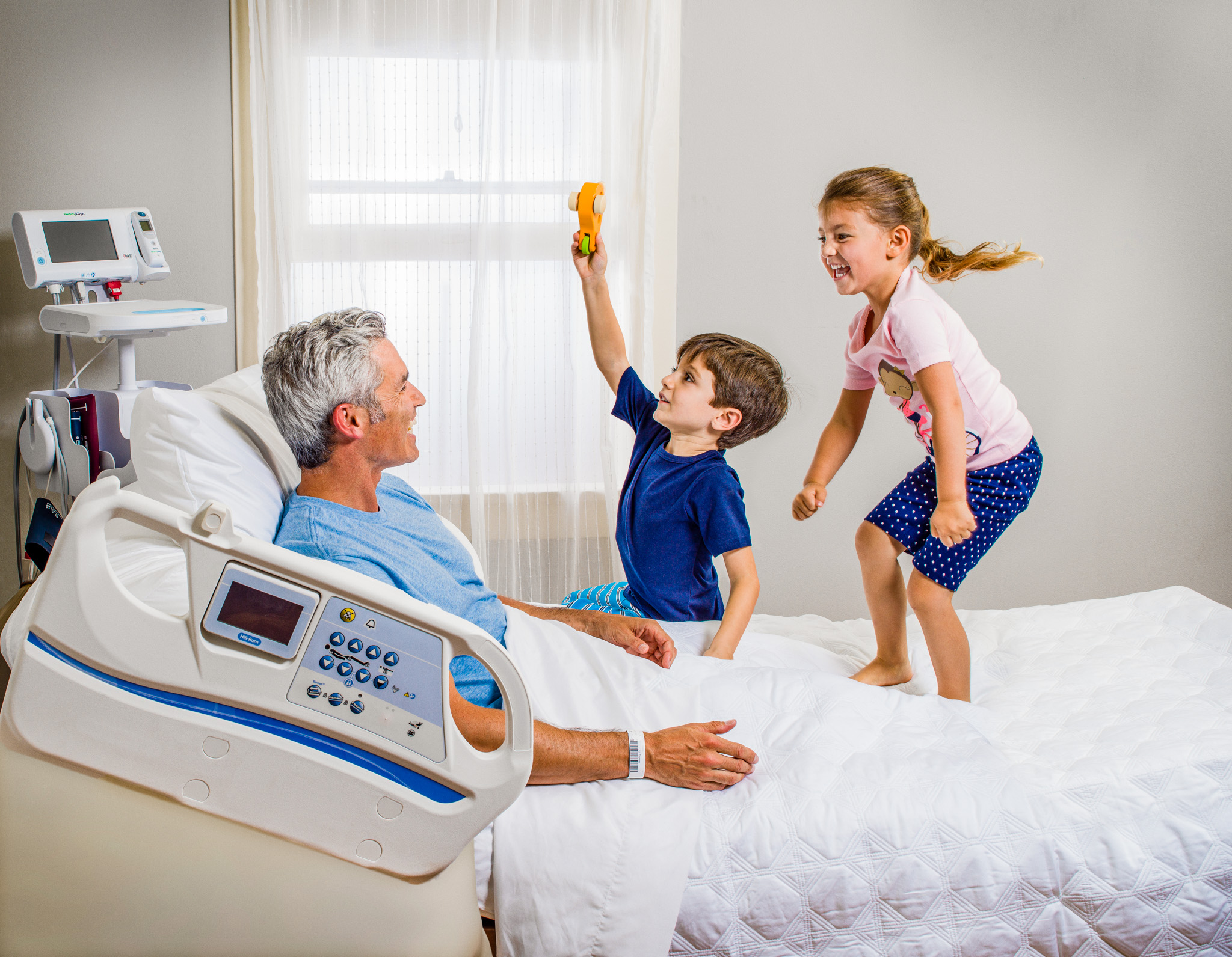 Photograph of a man at home in a hospital bed interacting with his children. © Dana Hursey Photography