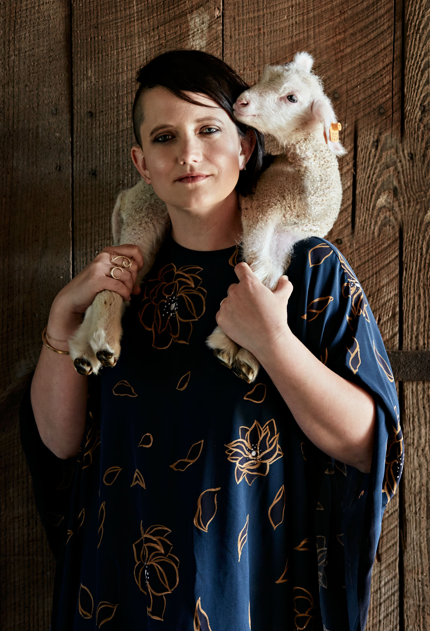 Editorial style portrait of hipster female farmer, wearing blue designer dress, with lamb draped around shoulders.