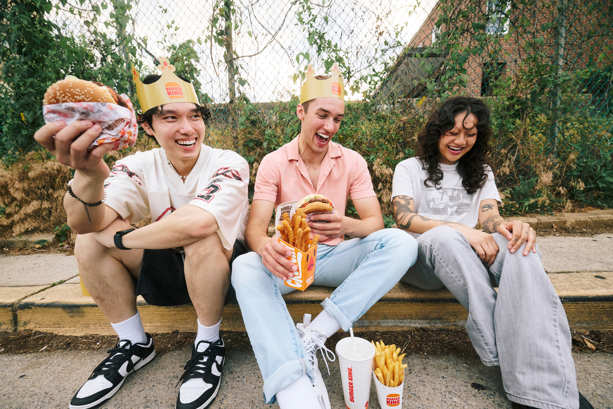 Friends laugh together while they eat Burger King on a sidewalk curb.