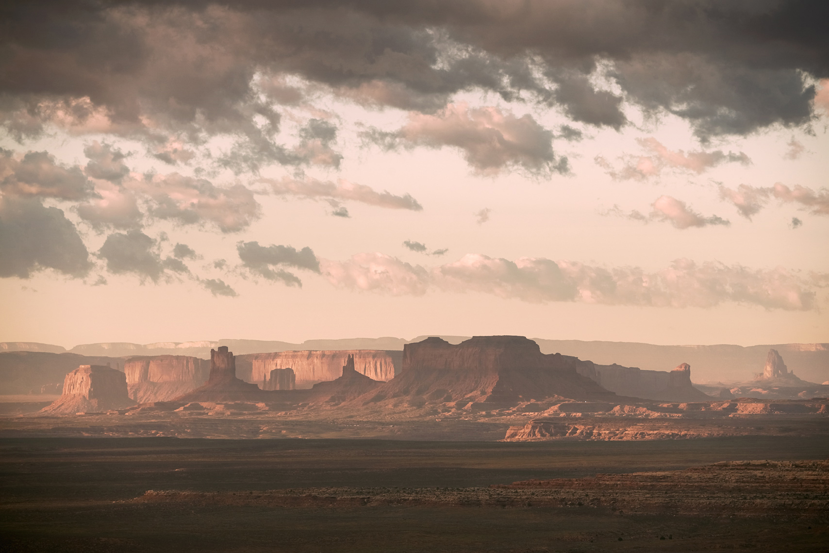 Low hanging clouds fill the sky above the picturesque landscape of Monument Valley in the four corners region of Southern Utah, Colorado, Arizona and New Mexico. Morning sunlight casts shows of the rock formations across the valley floor and up the rocky walls. 