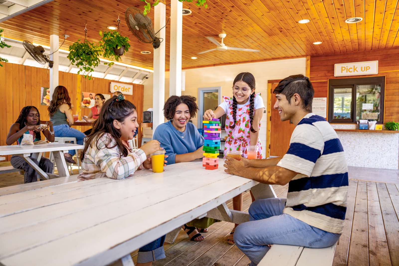 Family at a restaurant picnic table playing a stacking game with drinks, in a wooden café setting with plants and ceiling fans.