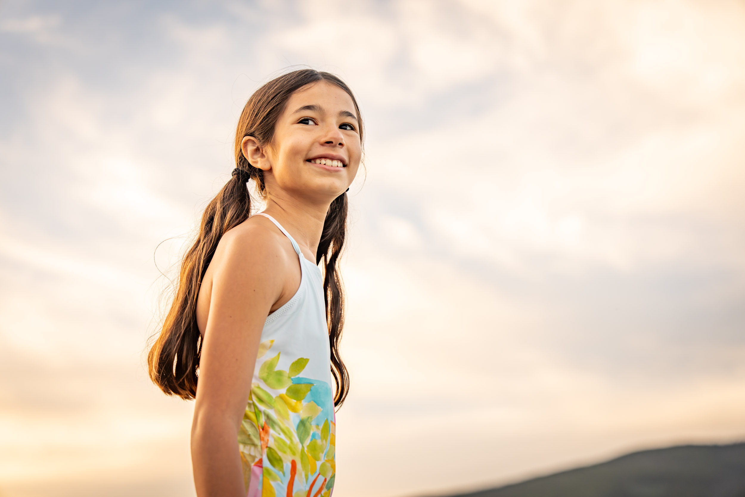 Young girl with long hair stands outdoors with a cloudy sunset sky and hills in the background.