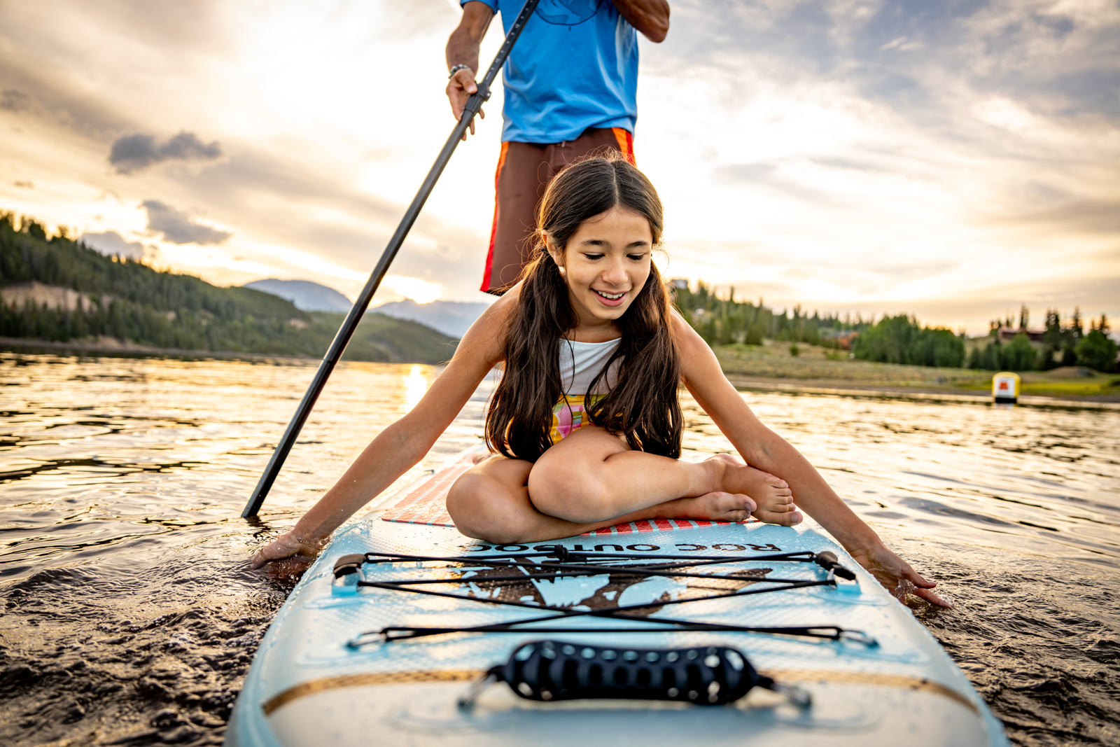 A girl sits on a paddleboard while a person stands and paddles on a lake with scenic mountains.