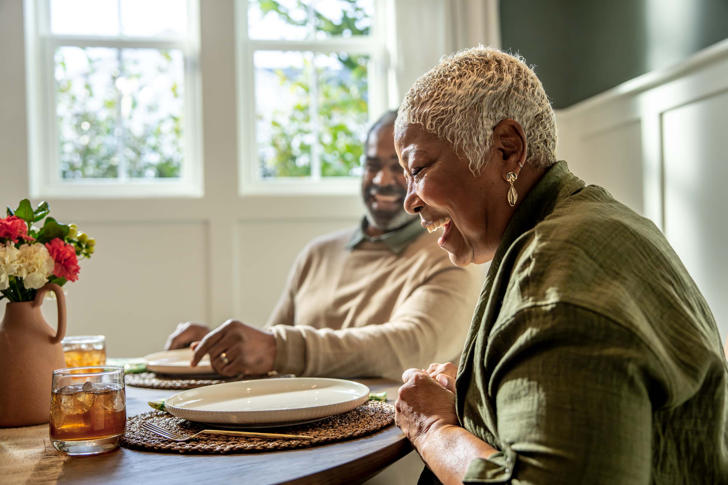 Mature couple sitting at a dining table with empty plates, a bright, cozy room. Woman is Laughing