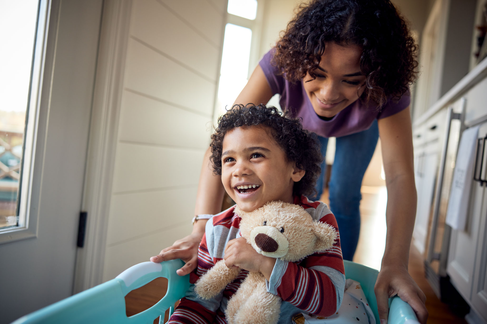 Older sister gives younger brother a ride in the laundry basket. African American teenage girl gives her younger brother a ride in a laundry basket. She pushes him around in a laundry basket with his stuffed bear on the hardwood floor of the home where they live.