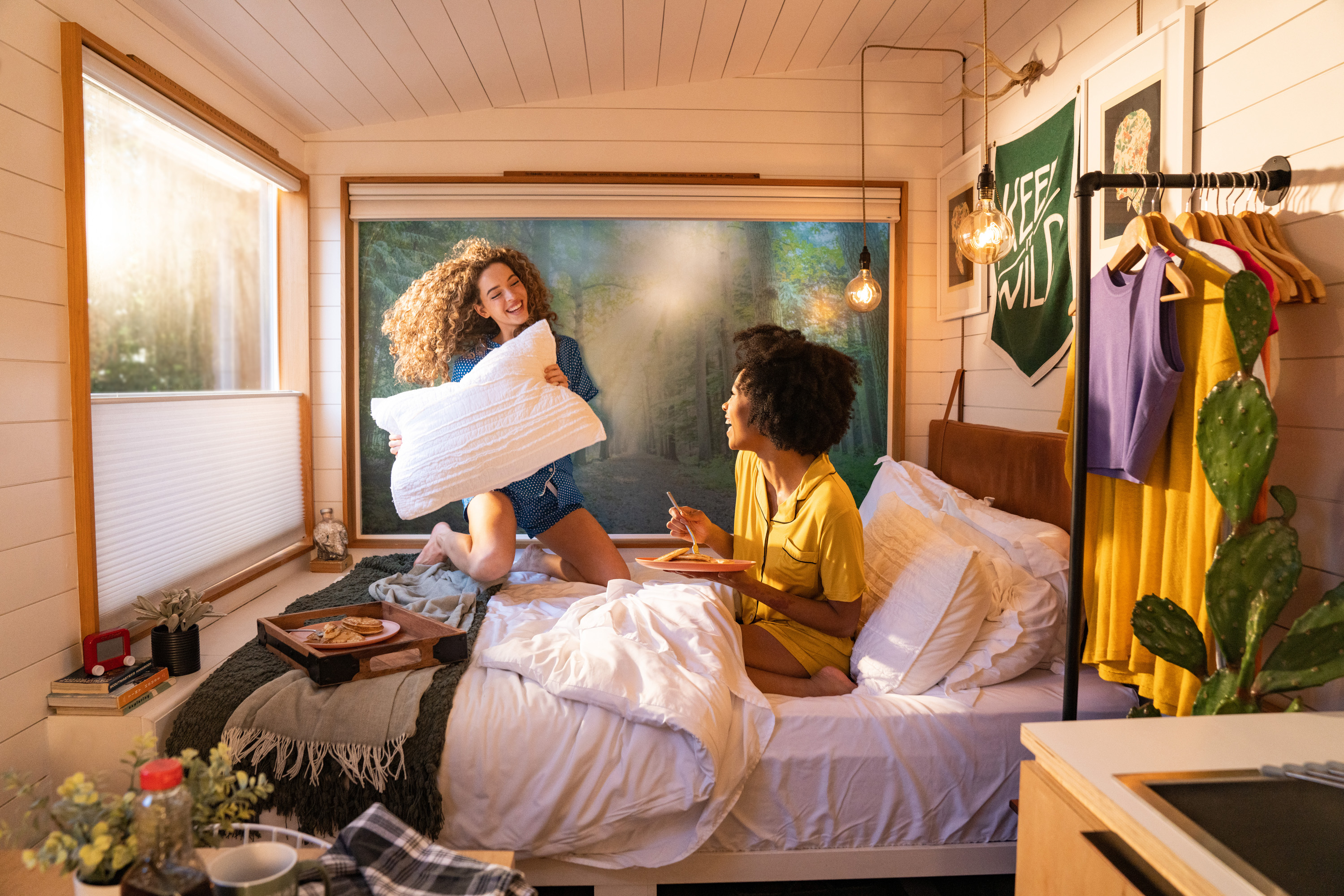 Lifestyle image of two girls having breakfast and jumping on the bed with morning sunshine through the windows.
