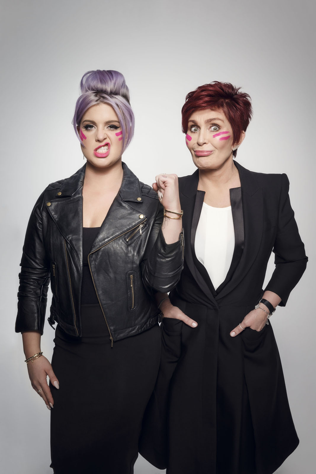 One woman wears a leather jacket and has purple hair, showing an angry expression. The other woman stands beside her in a black coat, relaxed but confident, both with unique makeup.