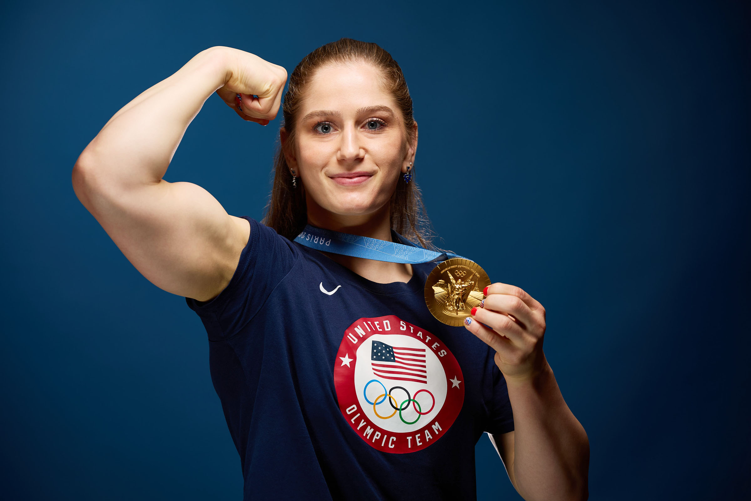 Athlete proudly displays gold medal while flexing bicep in studio setting. The scene captures dedication and achievement of Olympic level sports performance.