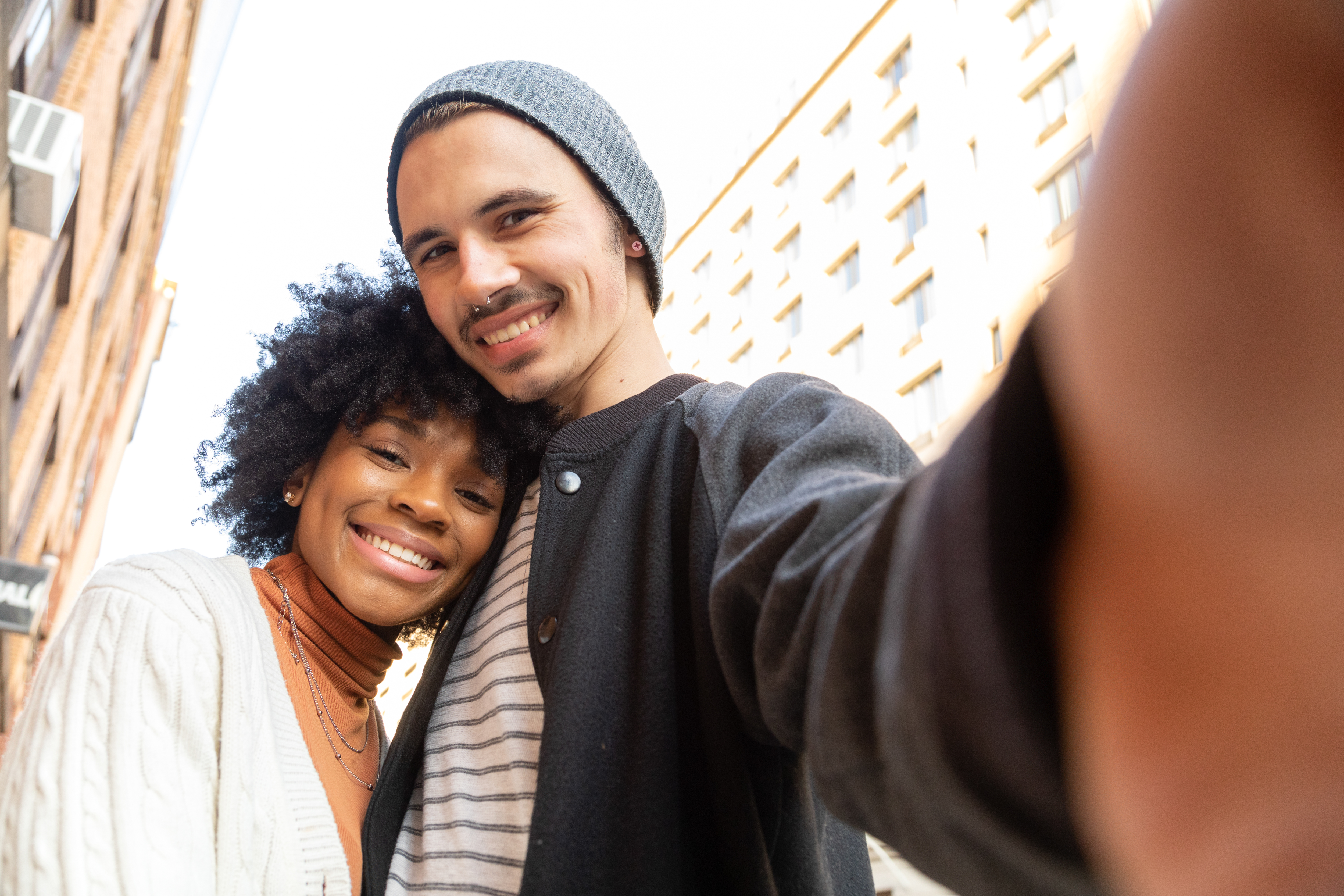 A young couple take a selfie together on a city street.  The man is caucasian, he has a nose ring and is wearing a grey hat, dark jacket, and white striped shirt with jeans.  The woman is African American and is wearing a white cardigan sweater and an orange turtleneck top with jeans, she has curly natural hair.  They are both in their early 20s and look like hipsters.  They are looking at the camera.  They are smiling.